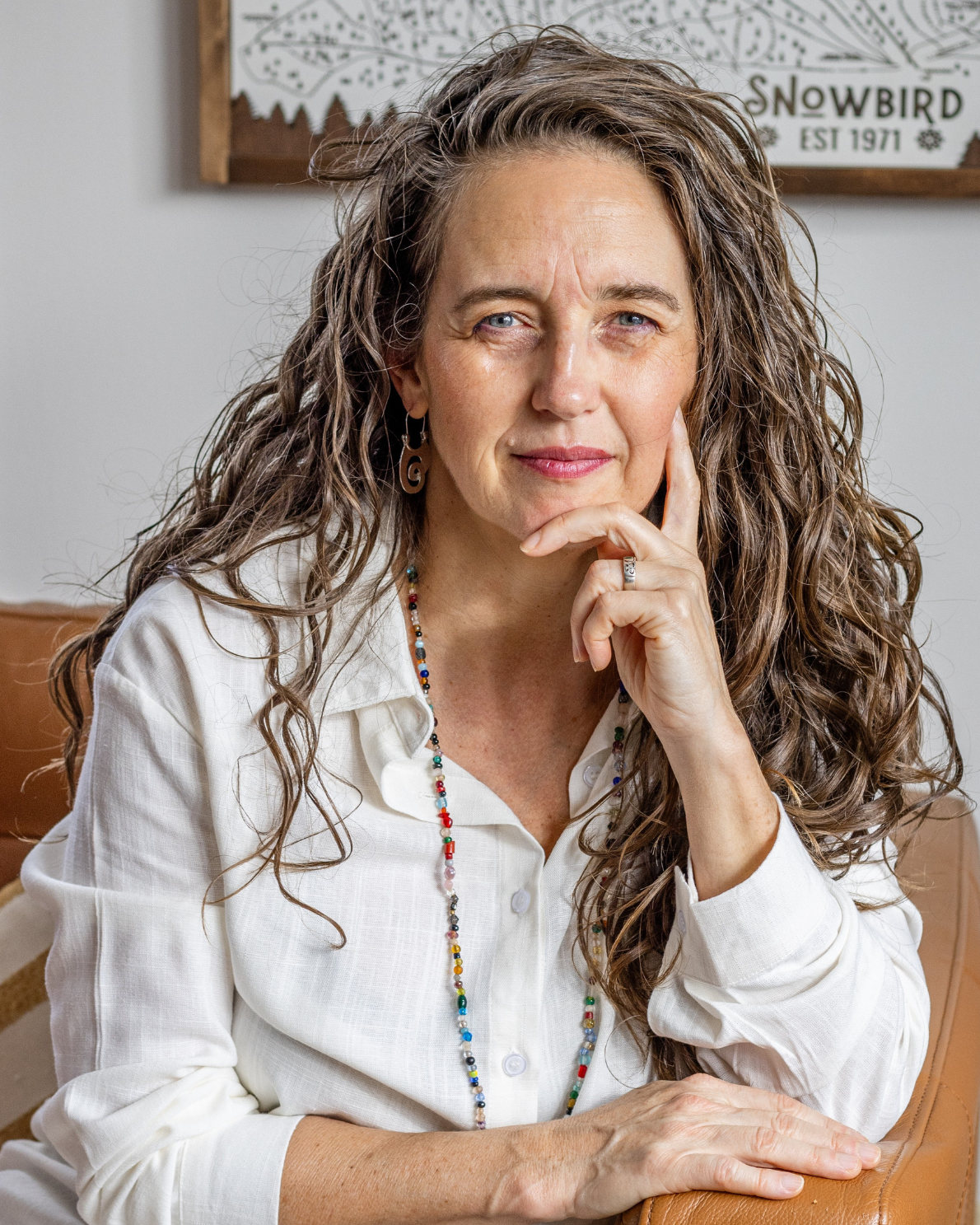Woman with long, wavy hair wearing a white shirt and colorful necklace, sitting indoors.